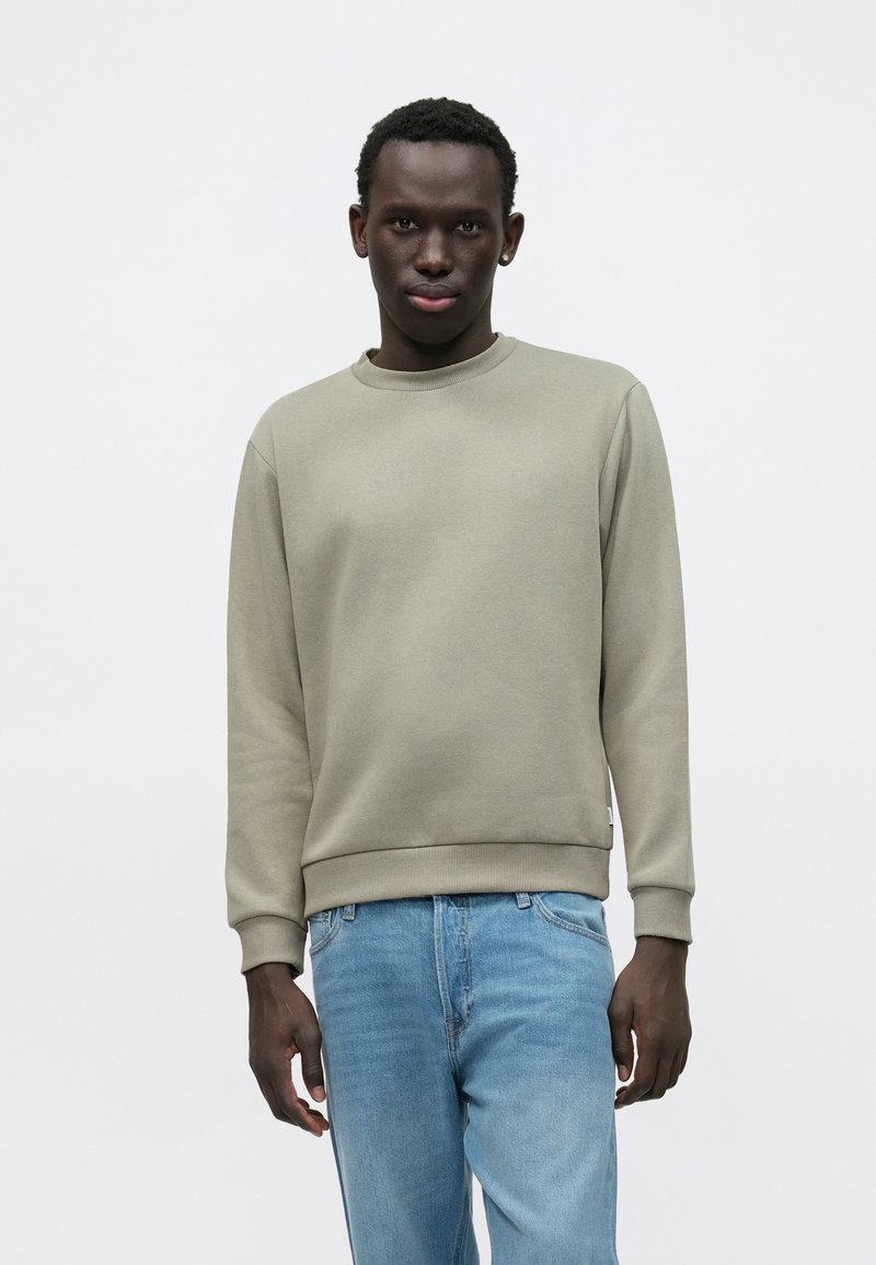 Felpa grigio chiaro con maniche lunghe, scollatura rotonda, polsini e orlo a coste. Abbinata a jeans azzurri, con una vestibilità comoda.