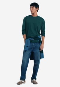 Grünes Langarmshirt, blauer Pullover um die Taille gebunden, blaue Jeans und graue Sneakers. Einfaches Design, lässige Passform und weiche Texturen.