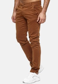 Brązowe, zwężane spodnie wykonane z miękkiego, gładkiego materiału, z klasycznym pięciopocketowym wzorem. Noszone z białymi butami sportowymi.