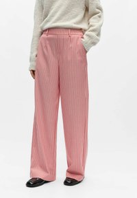 Rot-weiß gestreifte Hosen mit weitem Bein und plissiertem Taillendetail. Der Stoff wirkt glatt mit einer klaren Textur. Kombiniert mit einem hellfarbenen Pullover.