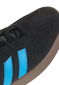 Schwarzer Wildleder-Sneaker mit zwei leuchtend blauen Lederstreifen an der Seite und dunkelbrauter Gummisohle, Nahaufnahme des Zehen- und Schnürbereichs.