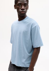 Giovane uomo che indossa una maglietta azzurro chiaro a maniche corte e jeans grigio scuro, in piedi con le mani in tasca, espressione neutra.
