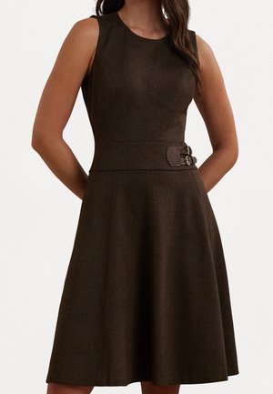 Bezrękawowa sukienka o długości do kolan w ciemnobrązowym kolorze, o dopasowanym kroju rozszerzającym się ku dołowi, z okrągłym dekoltem i podwójnym paskiem z klamrami w talii, noszona przez kobietę.