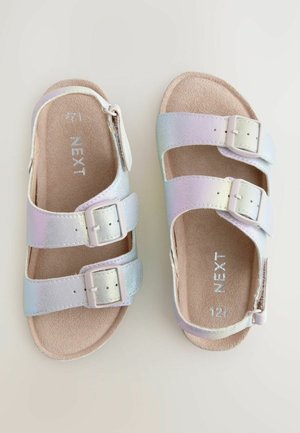 Iridescent čības ar diviem regulējamiem siksniņām, baltām sprādzēm un brūnu teksturētu pamatni. Iezīmējas ar gludu, metālisku apdari.