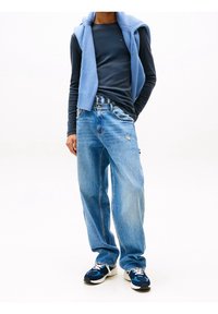 Lichtblauwe trui gedrapeerd over een donkerblauw shirt met lange mouwen. Draagt een versleten lichtblauwe spijkerbroek en donkerblauwe sneakers met witte zolen.