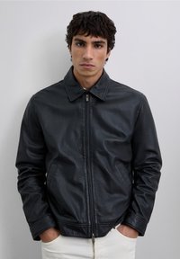 Joven con cabello rizado oscuro, vestido con una chaqueta de cuero negra con cremallera y pantalones blancos, manos en los bolsillos, expresión neutra, fondo simple.