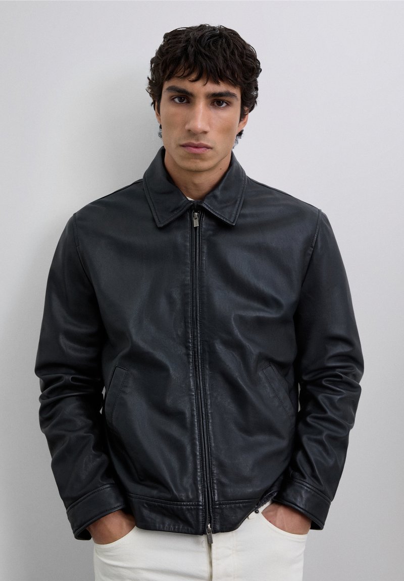 Joven con cabello rizado oscuro, vestido con una chaqueta de cuero negra con cremallera y pantalones blancos, manos en los bolsillos, expresión neutra, fondo simple.