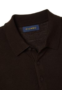 Polo marrone con colletto a coste, tre bottoni e una etichetta con la scritta "PIOMBO" in giallo su sfondo blu. Tessuto morbido.