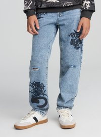 Jeans in denim azzurro chiaro con motivi graffiti e dettagli consumati, indossati con una felpa nera con stampa grafica e scarpe da ginnastica bianche con suola marrone.