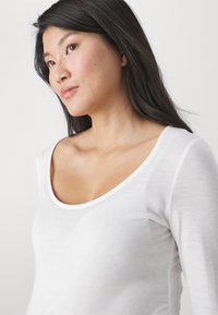 Blusa blanca de manga larga con un escote redondeado. Tela suave y ligera con una textura lisa, que presenta una silueta ajustada.
