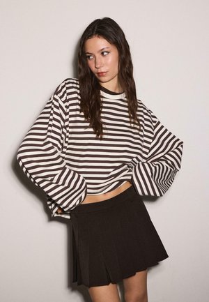 Mlada ženska z dolgimi temnimi lasmi nosi prevelik črno-bel pulover s črtami in črno nabrano mini krilo na enobarvni podlagi.