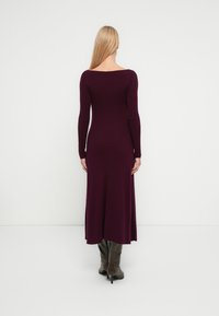 Langarmige, dieprode ribgebreide jurk met een open rug, nauwsluitend lijfje en een uitlopende zoom, gecombineerd met grijze laarzen tot aan de knie.