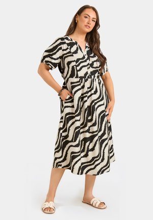 Femme debout, mains dans les poches, portant une robe midi à motif abstrait noir et blanc et des sandales blanches.