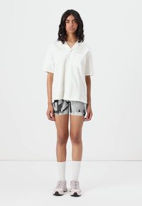 Camicetta bianca a maniche corte con taschino sul petto, abbinata a pantaloni corti con motivo bianco e nero e scarpe da ginnastica rosa chiaro con calzini bianchi.