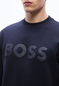 Άνδρας με σκούρο μπλε φούτερ που έχει μεγάλα ανάγλυφα γράμματα που σχηματίζουν τη λέξη "BOSS" στο στήθος.