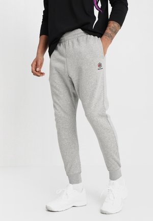 Mann trägt hellgraue Reebok-Sweatpants mit elastischen Bündchen, weiße Turnschuhe und ein schwarzes Langarmshirt mit lila Design.