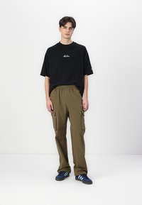 Black cotton t-shirt with white logo, paired with olive green cargo pants featuring side pockets, completed with blue sneakers and stripes.