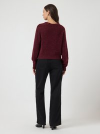Mulher de costas, vestindo um suéter felpudo borgonha, calças largas pretas e botas de cano curto com salto pretas, em um fundo branco liso.