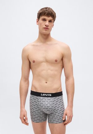 Νεαρός άνδρας που στέκεται με ουδέτερη έκφραση, φορώντας γκρι εσώρουχα μπόξερ της μάρκας Levi's με μαύρη ζώνη μέσης.