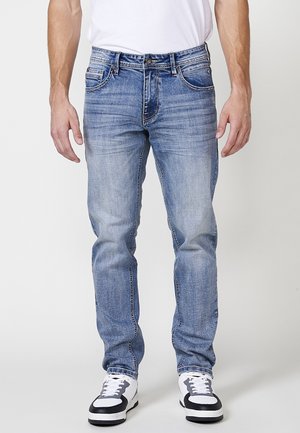 Jeans de denim azul claro con corte recto, acabado desgastado, cinco bolsillos y cierre de botón. Combinados con zapatillas blancas con detalles en negro.