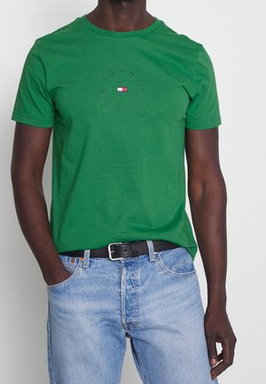 Personne portant un t-shirt vert manches courtes avec un logo circulaire et un jean bleu clair avec ceinture noire, une main dans la poche, sur fond blanc.