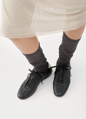 Άτομο που φοράει μαύρα αθλητικά παπούτσια και διάφανες μαύρες κάλτσες, στέκεται σε λευκό πάτωμα με φούστα ανοιχτού μπεζ χρώματος που φαίνεται πάνω από τα γόνατα.