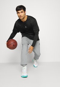 Černé sportovní tričko s dlouhým rukávem, šedé tepláky a bílé tenisky s tyrkysovými akcenty. Hráč drž ící basketbal s logem.