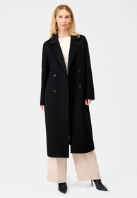 Sort uld dobbeltbrystet frakke med store knapper, spids krave, lange ærmer og lige snit, båret over beige brede bukser.