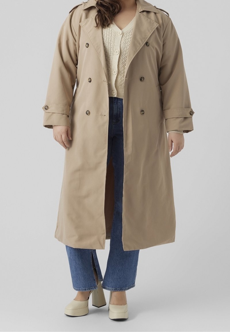 Beige trenchcoat met dubbelzijdige knopen en grote revers. Draag het over een crème trui en blauwe high-waisted jeans. Stevige, lichtgekleurde hakken.