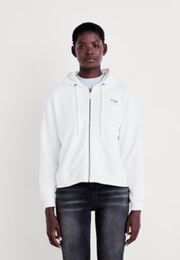 White zip-up hoodie with a drawstring hood and subtle logo in pastel colors on the chest. Soft fabric, cropped design, paired with dark jeans.