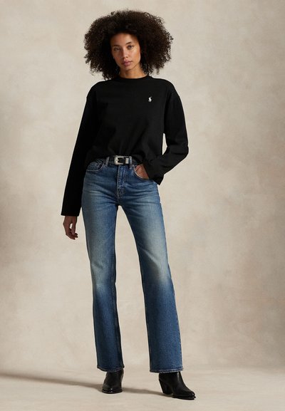 Kobieta stoi z ręką w kieszeni, ubrana w czarną koszulę z długim rękawem, wyblakłe niebieskie jeansy z wysokim stanem, czarny pasek i czarne botki na obcasie.