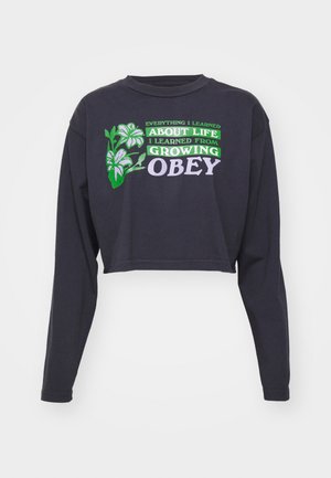 Zwarte cropped longsleeve met groene en paarse tekst "Alles wat ik heb geleerd... over het leven heb ik geleerd van het groeien OBEY," met bloemenaccenten.