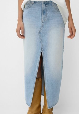 Jupe midi en denim bleu clair avec fente centrale à l'avant, portée avec des bottes hauteur genou couleur camel et un top blanc.