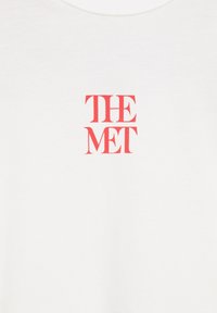 Tissu blanc avec texte serif rouge « THE MET » centré sur la partie haute de la poitrine, probablement une partie d’un vêtement tel qu’un t-shirt.