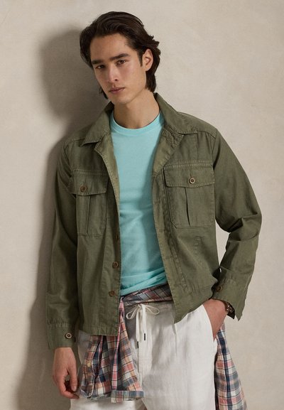 Νέος άνδρας που φοράει ένα ελαιί πράσινο μπουφάν πάνω από ένα ανοιχτό μπλε πουκάμισο, με ένα καρό πουκάμισο δεμένο γύρω από τη μέση του και λευκό παντελόνι, στέκεται μέσα σε εσωτερικό χώρο.