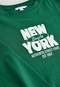 Zeleni pamučni T-shirt s okruglim izrezom. Ima bijeli otisnuti tekst: "NEW YORK Bagels AUTENTIČNA ULICA HRANA OSNIVAJU 1997." Glatka tekstura.