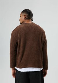 Bruine, gestructureerde, oversized trui met lange mouwen en een ronde halslijn. Gedragen over een wit overhemd, gezien van achteren.