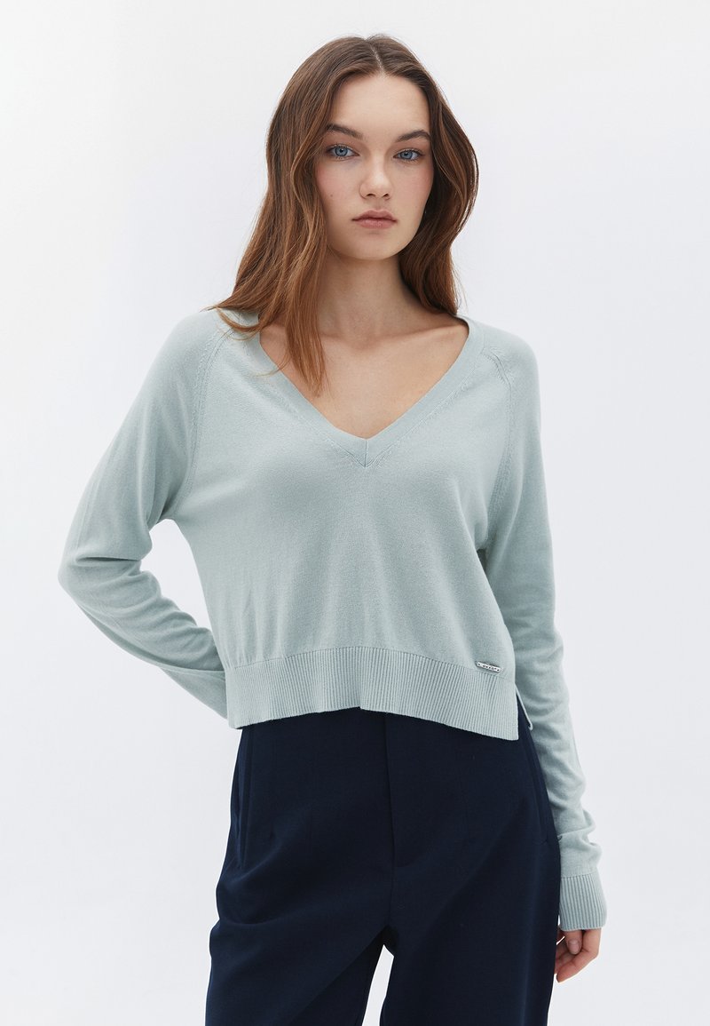 Suéter corto verde claro con escote en V y mangas largas; presenta un dobladillo acanalado y un sutil detalle de costura. Combinado con pantalones oscuros.