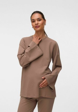 Femme aux cheveux châtains clairs attachés en arrière porte un haut marron ample à manches longues et un pantalon, une main sur la poitrine et l'autre sur le ventre.