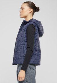 Mörkblå quiltad väst med huva, med dragkedja fram och utan ärmar. Kombinerad med en svart långärmad tröja och blå denimjeans.