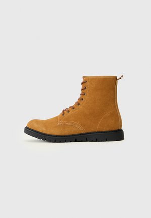 Bottine en suède beige avec un bout rond, des lacets marron, un talon renforcé et une semelle en caoutchouc noire. Elle présente des accents de couture et une boucle à l'arrière.