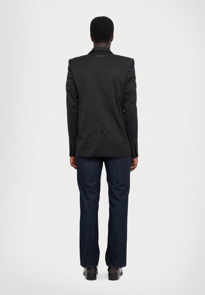 Blazer noir avec épaules structurées, fente simple, associé à un jean droit bleu foncé et des chaussures noires, vu de dos.