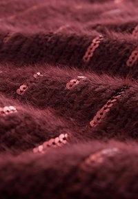 Bordeaux gestructureerde stof met een fuzzy uitstraling, voorzien van rijen glanzende roze pailletten die op verschillende plekken zijn geplaatst.
