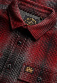 Camisa de cuadros rojos y negros abotonada, con cuello y etiquetas de la marca Superdry en el interior del cuello y del bolsillo del pecho.