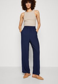 Pantalons larges bleu marine avec une taille élastique et des poches latérales, associés à un haut sans manches beige et des chaussures tan à enfiler.