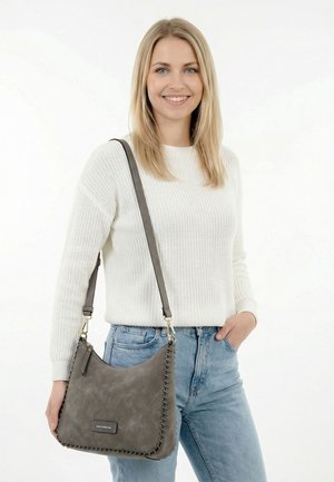 Hymyilevä nainen, joka pukeutuu valkoiseen neulepuseroon ja vaaleisiin farkkuihin, pitää käsissään harmaata nahkaista käsilaukkua, jossa on tikatut reunat ja olkahihna.