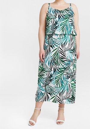 Femme portant une robe sans manches, longue jusqu'aux chevilles, avec des motifs de feuilles tropicales vertes, bleues et noires, et des talons blancs à bride cheville.