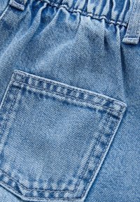 Tissu en denim bleu clair présentant une taille froncée et une petite poche à patch avec des détails cousus. Surface texturée avec une légère décoloration.