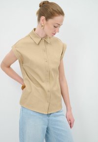 Camicia beige a maniche corte con colletto classico, caratterizzata da una superficie liscia e un'orlo arrotondato, abbinata a jeans azzurri chiari.