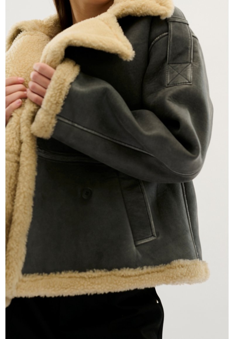Chaqueta gris forrada de piel de oveja con un suave cuello y borde en crema; cuenta con cierre de doble botón y bolsillos laterales. Textura suave y diseño en capas.